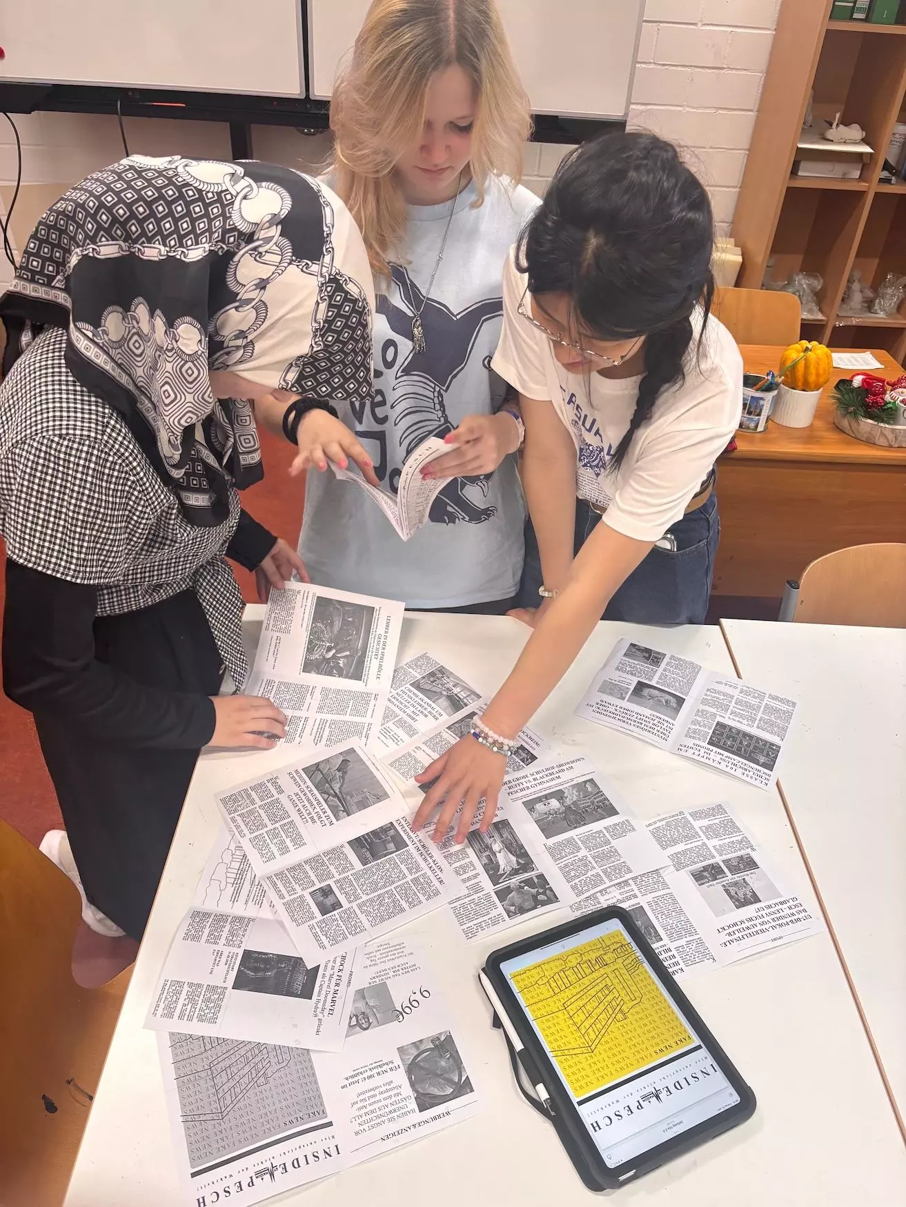 Schülerinnen stehen vor einem Tisch und schauen auf die einzelnen Seiten der Fake News Zeitung, die sie gebastelt haben.