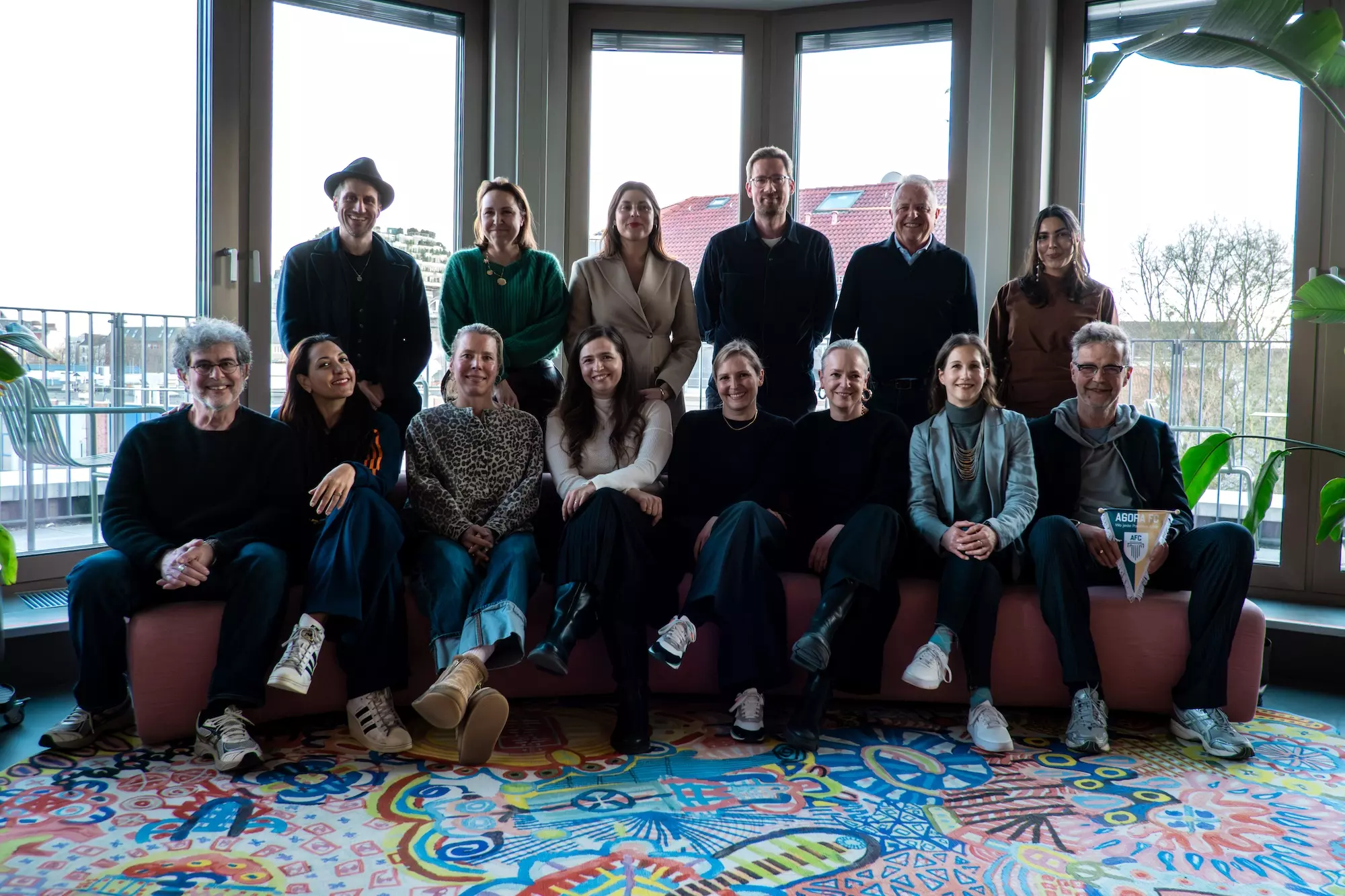 ProjektZUVERSICHT_Kreativwettbewerb_Jury: 14 Personen stehen bzw. sitzen vor einem Fenster.