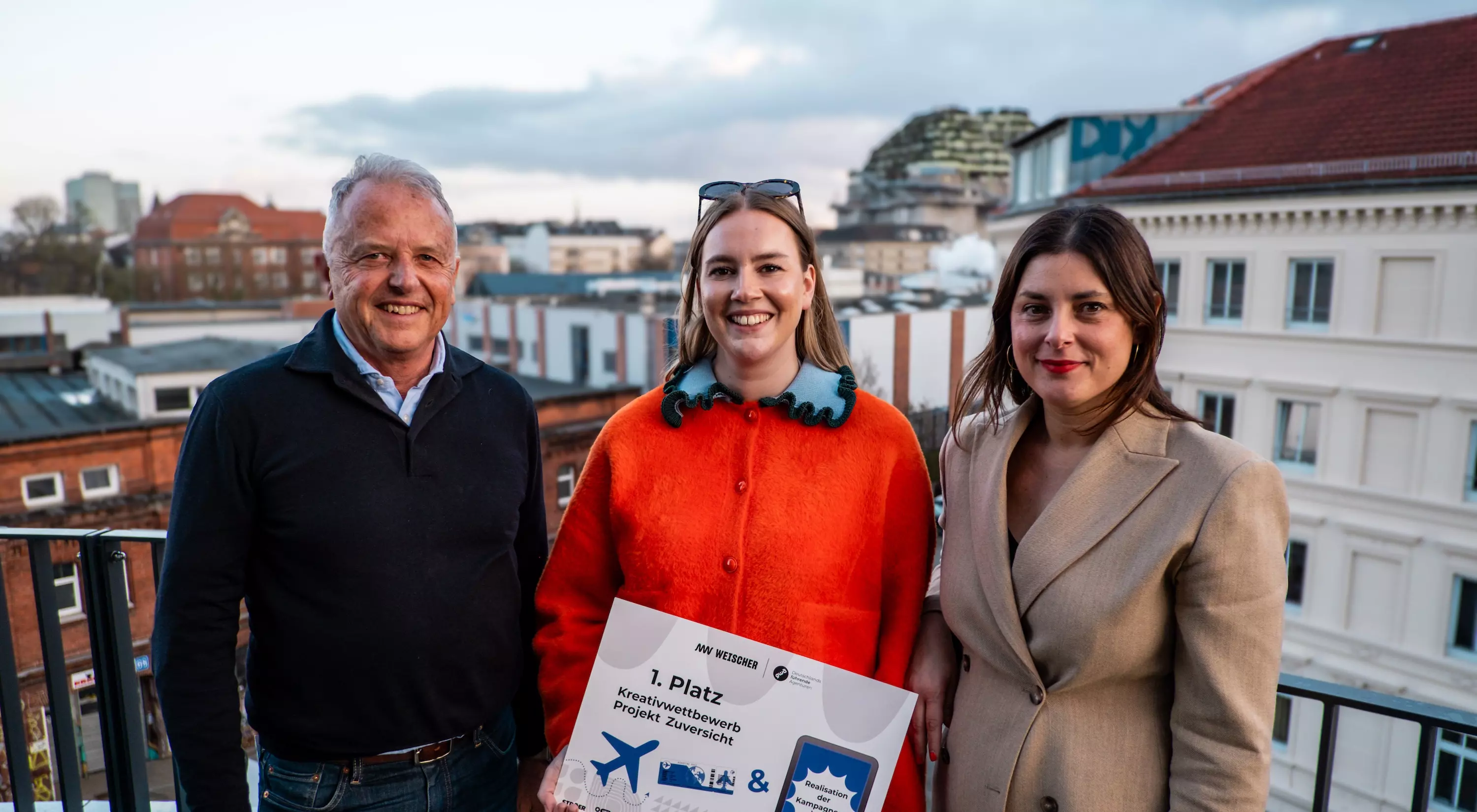 Eine der Siegerinnen vom Kreativwettbewerb zum Proiekt Zuversicht steht mit Florian Weischer von Weischer und Claudia Díaz Sánchez von GWA auf einem Balkon und lächelt.
